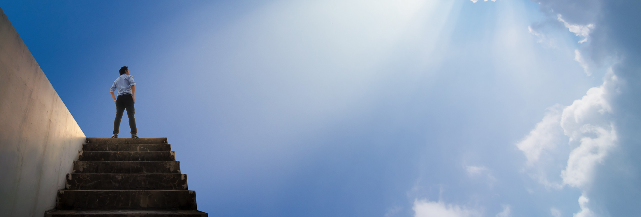 A man stands at the top of a staircase, looking up towards a bright blue sky with rays of sunlight and clouds.