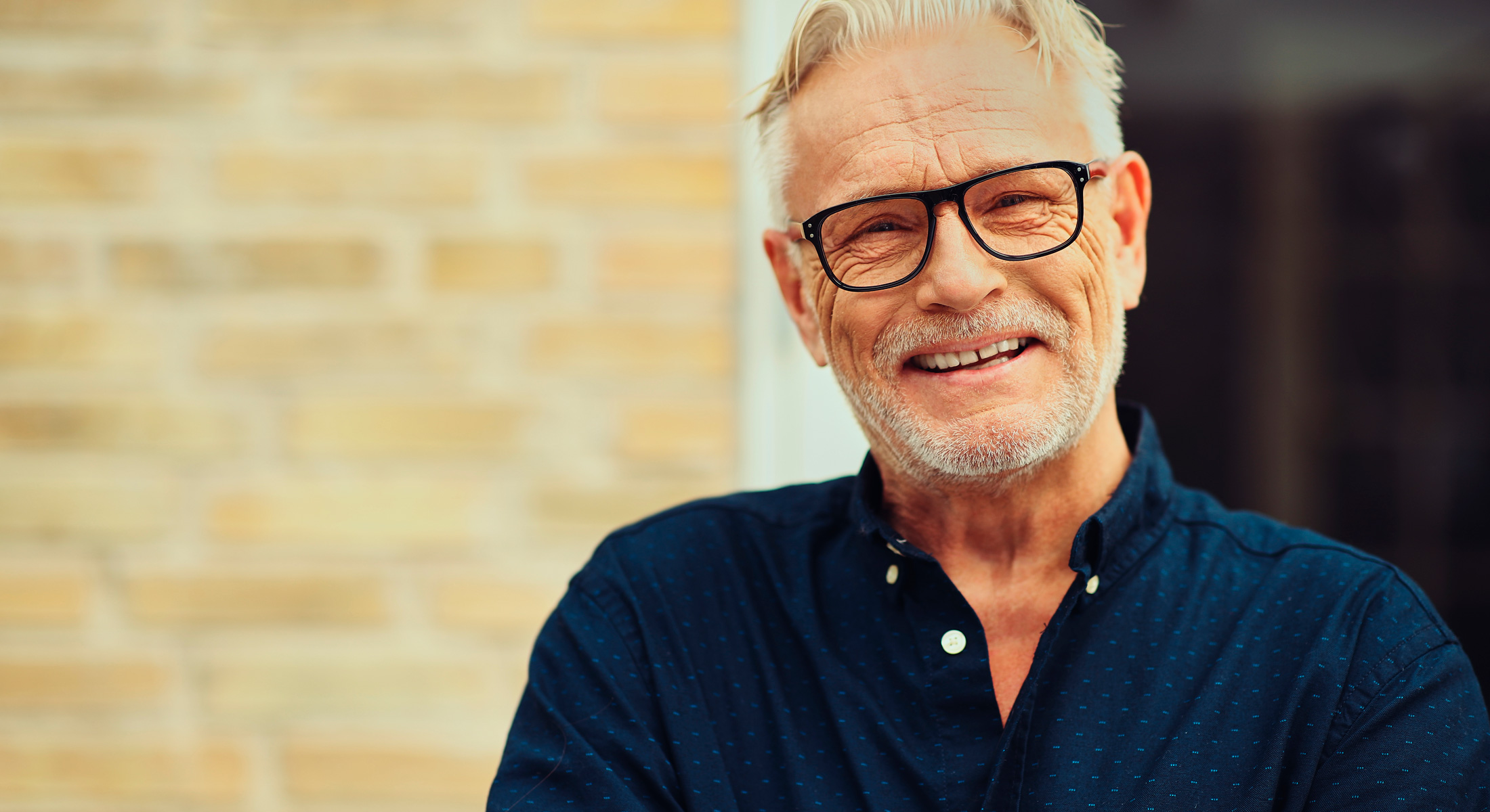 A mature man with gray hair and glasses smiles warmly at the camera, standing in front of a brick wall.