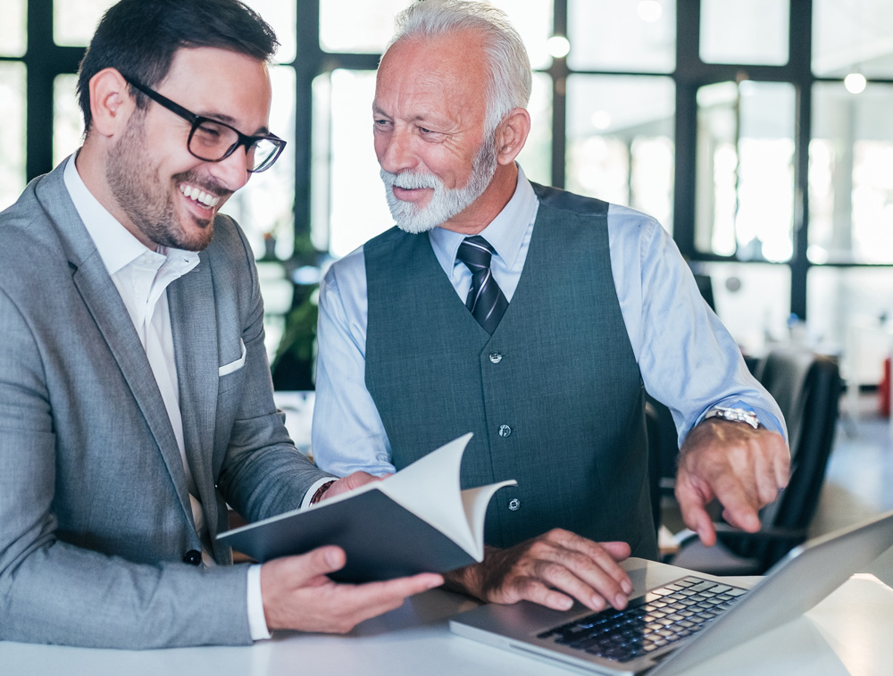 A businessman and a senior professional engaging in discussion over a laptop, with one holding a book and both sharing smiles in a modern office environment.