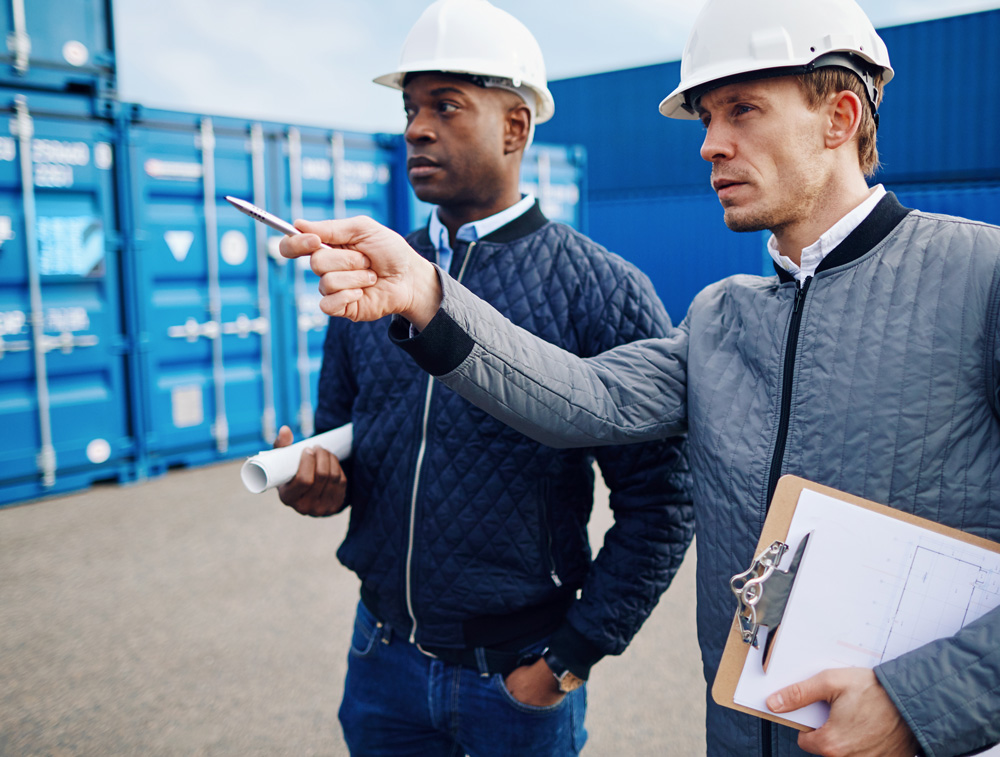 Two construction professionals wearing helmets, one pointing and discussing while holding blueprints, with shipping containers in the background.