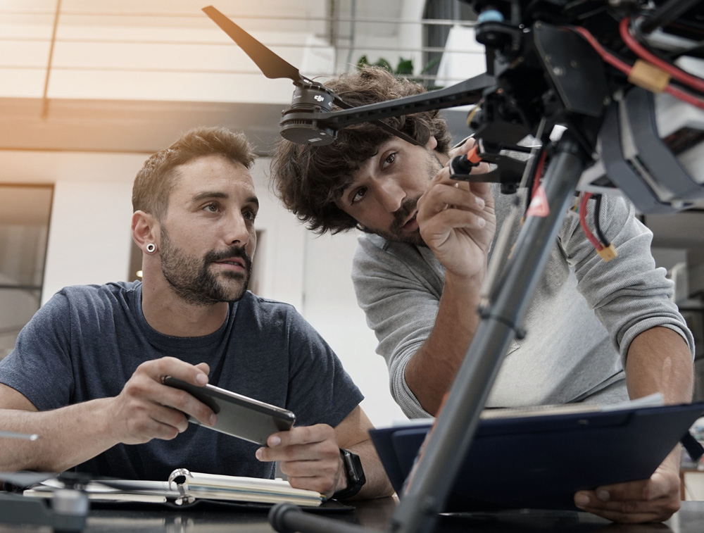 Two men working together on a drone, one holding a smartphone and the other inspecting the drone's components.