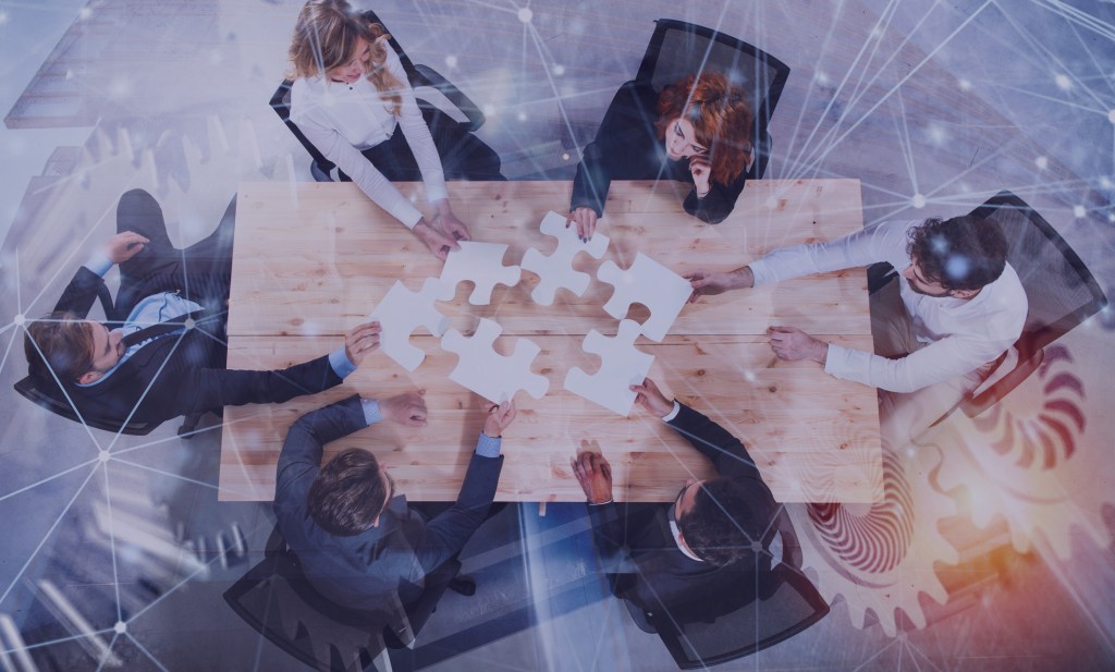 A group of six professionals collaborating at a wooden table, working together with puzzle pieces, symbolizing teamwork and problem-solving.