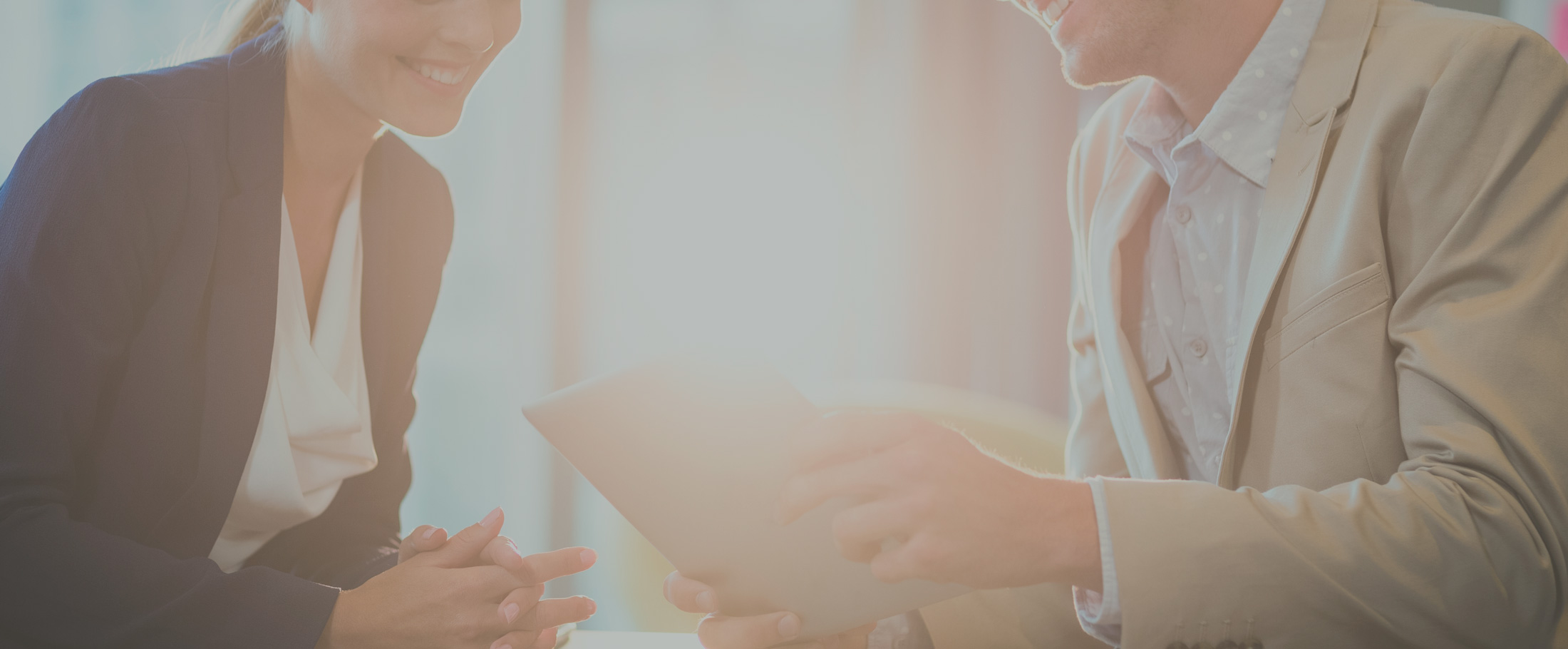 A close-up of two professionals, a man and a woman, engaged in a discussion, smiling while looking at a tablet in a bright office setting.