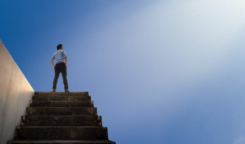 A person standing at the top of a set of stairs, looking up towards a bright blue sky.